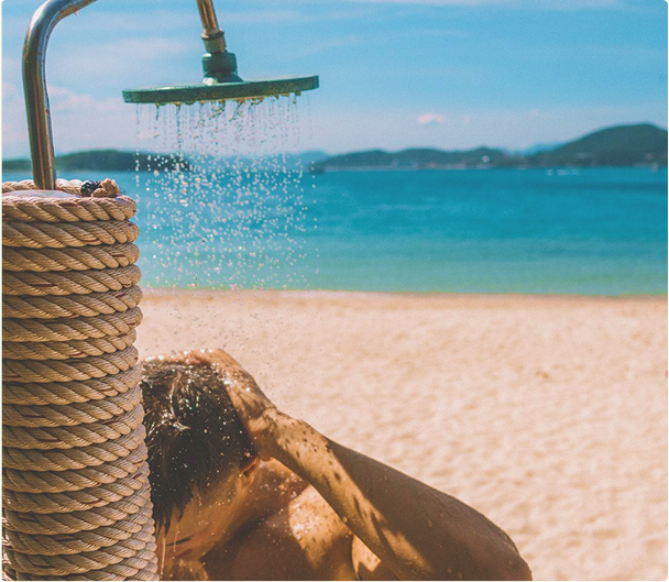Person taking an outdoor shower on a beach with ocean and sky in the background before applying post surf serum